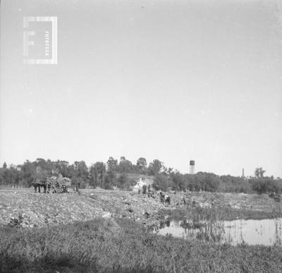 Basural con gente, carro y barranca con tanque Aguas Corrientes al fondo