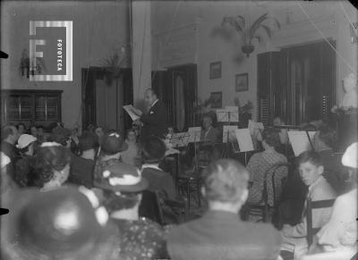 Sr. leyendo, orquesta atrás, público en 1er. plano